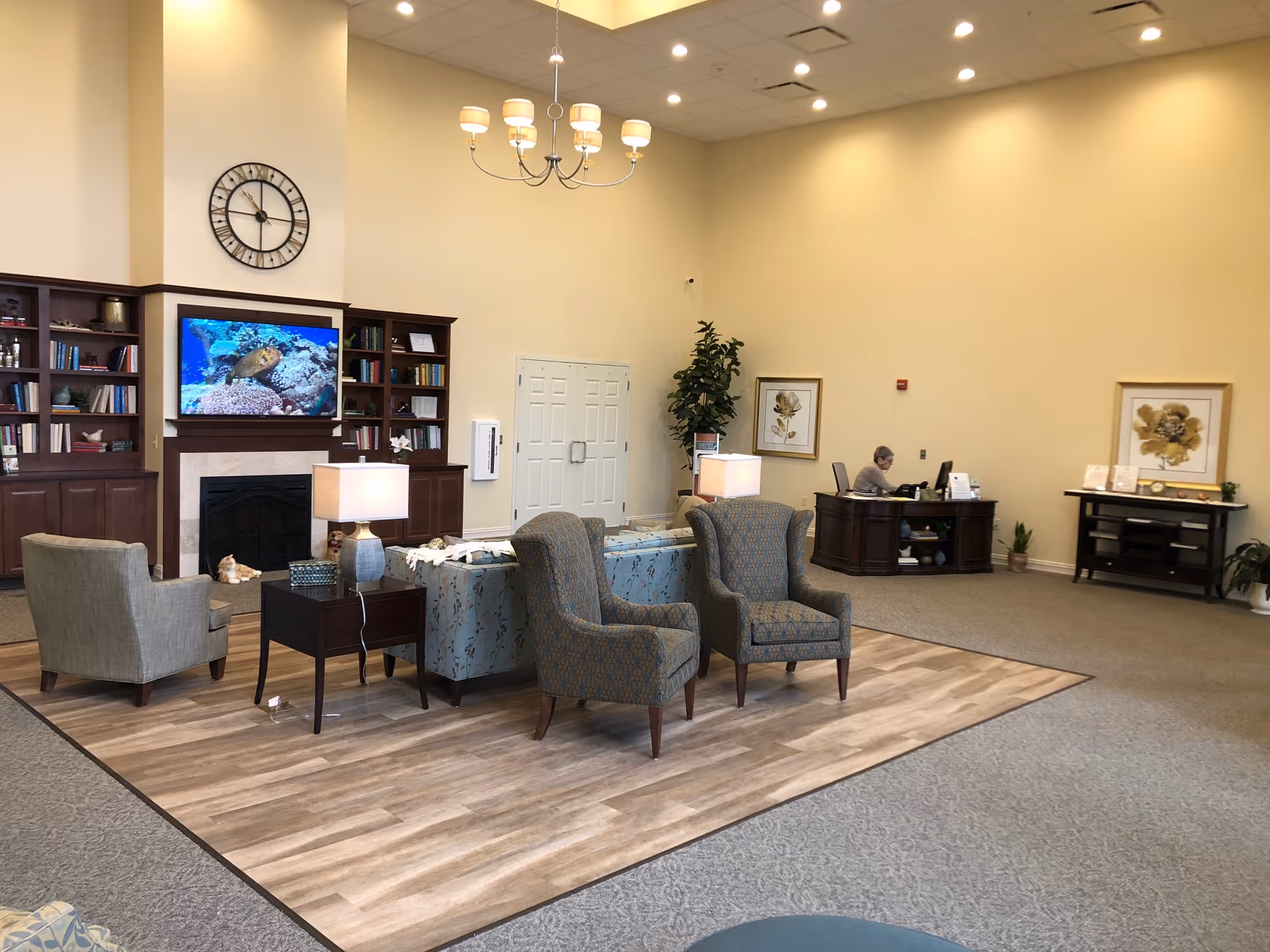 A spacious senior living facility common area with high ceilings and warm lighting. The room features a seating arrangement with two patterned armchairs, a blue floral sofa, and a gray armchair around a wooden side table with a lamp. Behind the seating area is a fireplace with a mounted TV displaying an underwater scene with a sea turtle. Built-in wooden bookshelves flank the fireplace, filled with books and decorative items. A large clock hangs above the fireplace. In the background, a person is seated at a wooden reception desk, and there are framed floral artworks on the walls. The floor is a combination of wood and carpet.