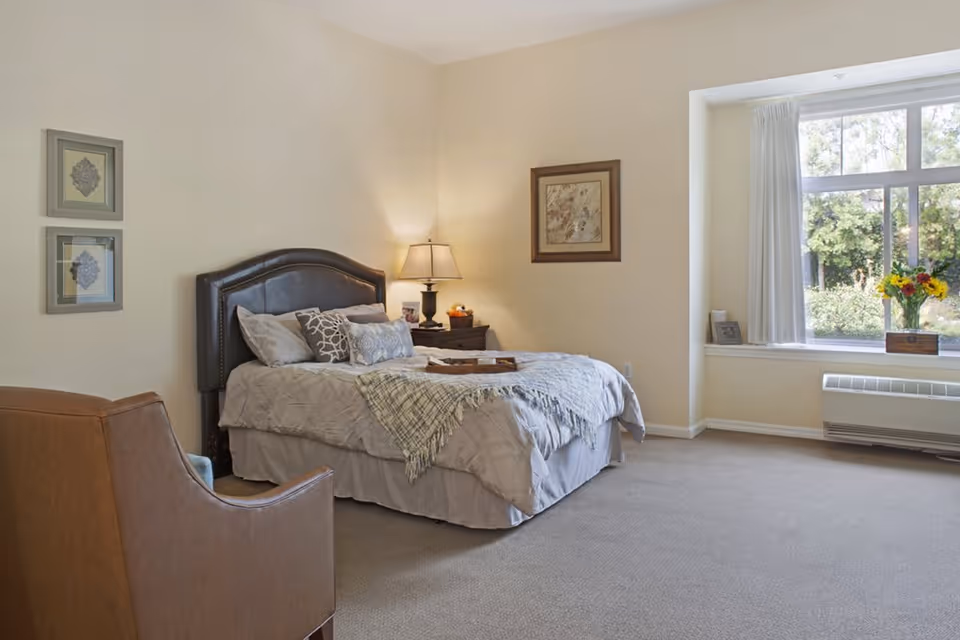 A cozy bedroom with a neatly made bed featuring multiple pillows and a throw blanket. There is a brown leather armchair in the foreground, a nightstand with a lamp and decorative items beside the bed, and framed artwork on the walls. A large window with white curtains lets in natural light and offers a view of greenery outside.