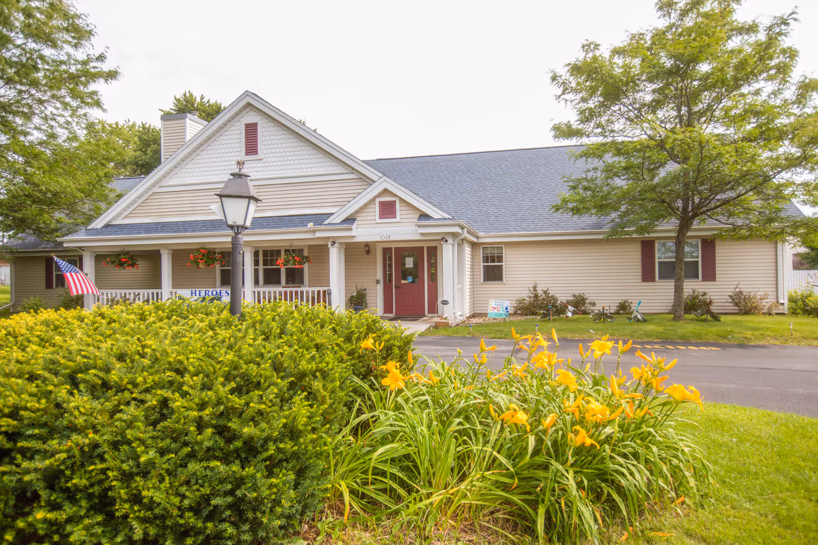 Front exterior of a single-story assisted living building with a porch, lamp post, American flag and yellow flowers in the foreground.