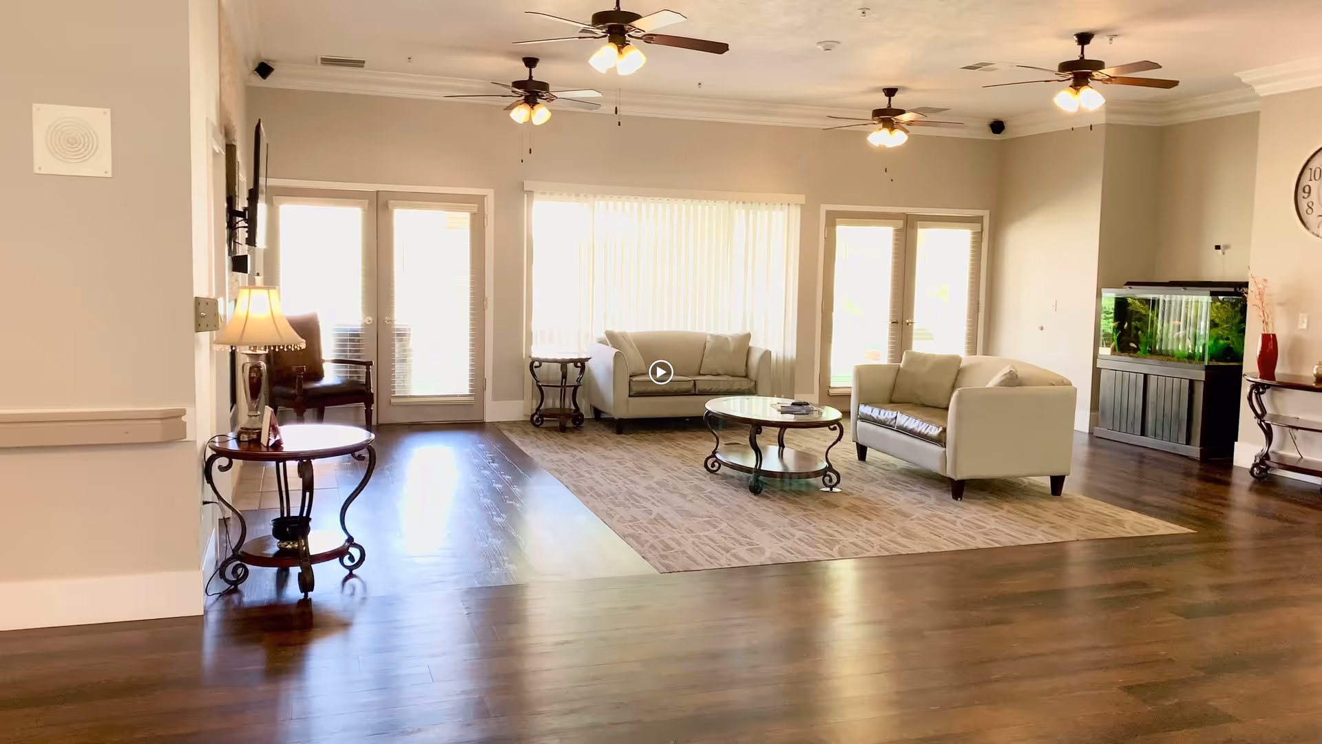 A bright and spacious living room area with two beige sofas arranged around a glass coffee table on a patterned rug. The room features hardwood floors, ceiling fans with lights, a large fish tank on a stand, a wall clock, and multiple windows and glass doors allowing natural light to fill the space. There are also side tables with lamps and chairs near the windows.