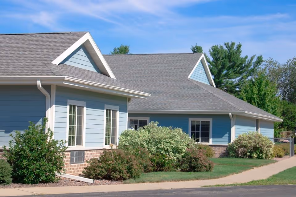 Exterior view of a single-story building with blue siding, white trim, and a gray shingled roof. The building is surrounded by green shrubs and a sidewalk, with trees and a blue sky in the background.