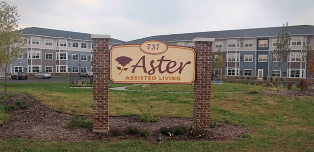 Sign reading "Aster Assisted Living 737" mounted between brick pillars on a lawn with a multi-story assisted living building in the background.