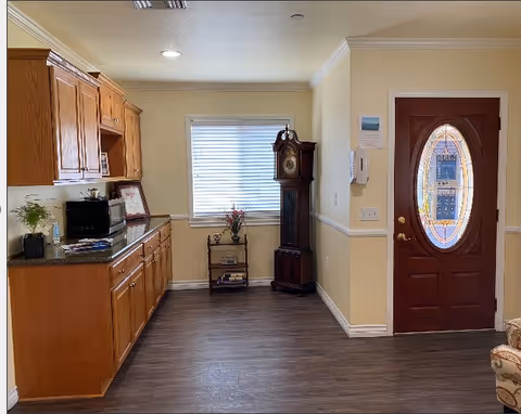 Interior view of a room with wooden cabinets and a countertop on the left, a window with blinds in the center, a tall grandfather clock next to a small shelf with decorative items, and a wooden door with an oval glass panel on the right.