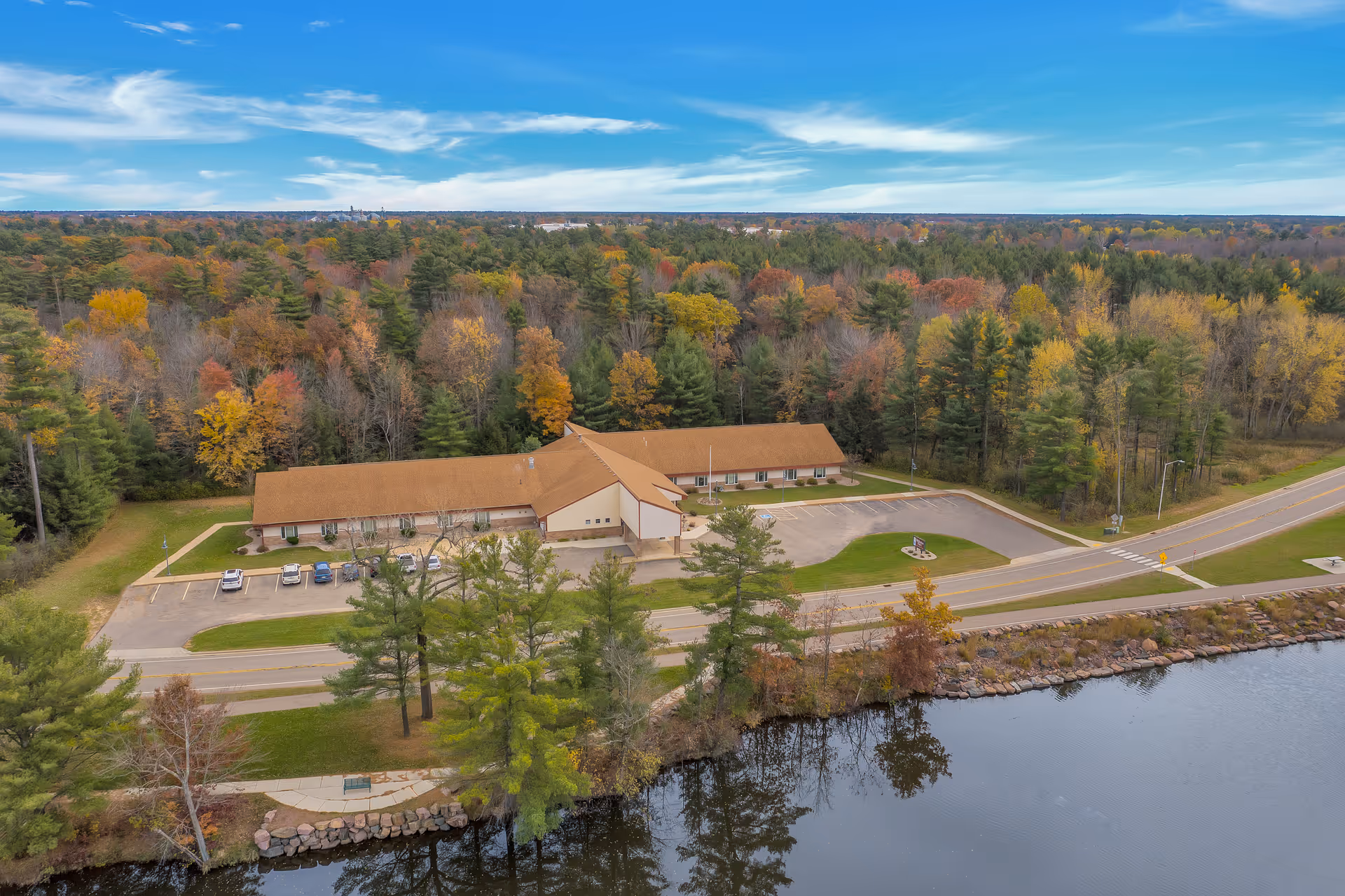 Aerial view of Wellington Place at Biron, a single-story building with a brown roof surrounded by trees with autumn foliage. The building is adjacent to a road and a body of water, with a parking lot containing several cars in front.