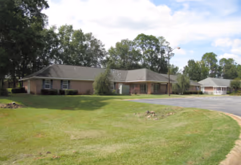 Single-story brick building with a gray shingled roof surrounded by green grass and trees under a partly cloudy sky.