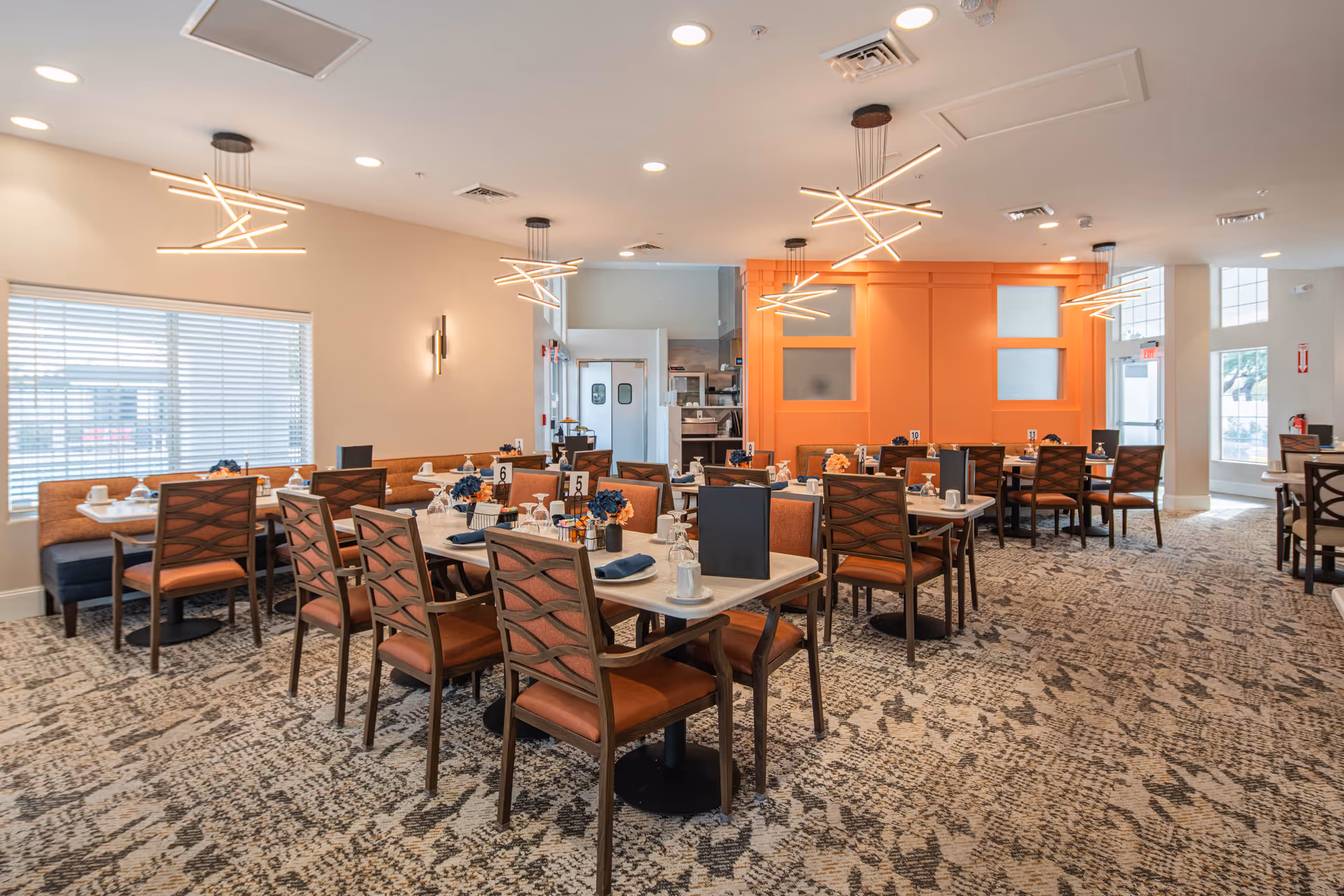 A bright, modern dining room with multiple set tables, orange accent wall, and contemporary pendant lights.