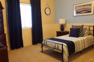 A tidy bedroom with a single metal-framed bed dressed in striped bedding and navy accents, a nightstand with lamp, dresser, wall clock, window with dark curtains, and framed art.