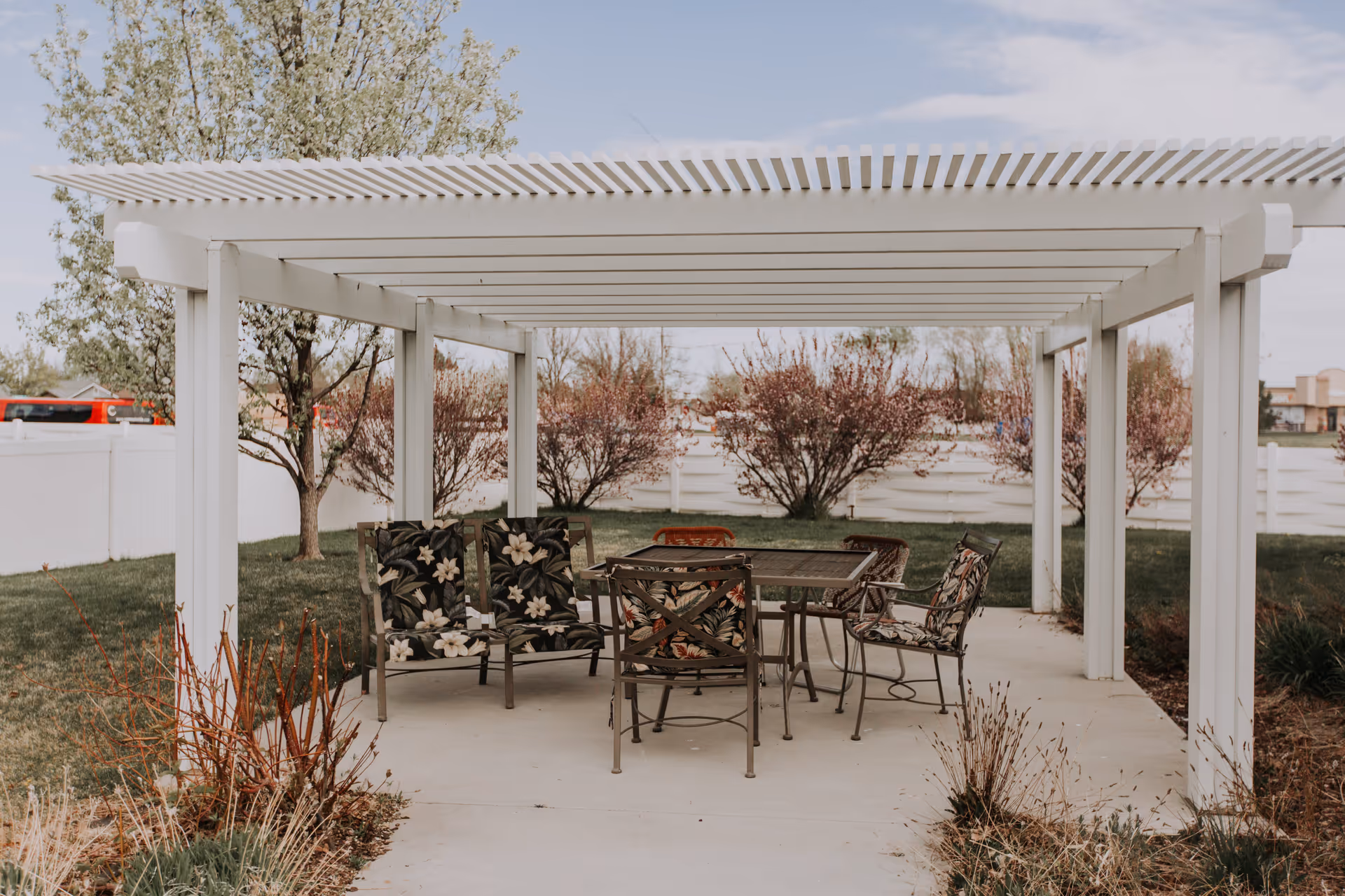 Outdoor patio area with a white pergola covering a set of metal chairs and a table. The chairs have floral patterned cushions. Surrounding the patio are green grass, bushes, and trees with some pink blossoms. A white fence encloses the area.