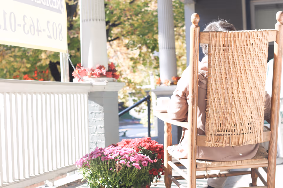 An older person sits in a wicker rocking chair on a sunny porch with potted pink flowers and white columns.