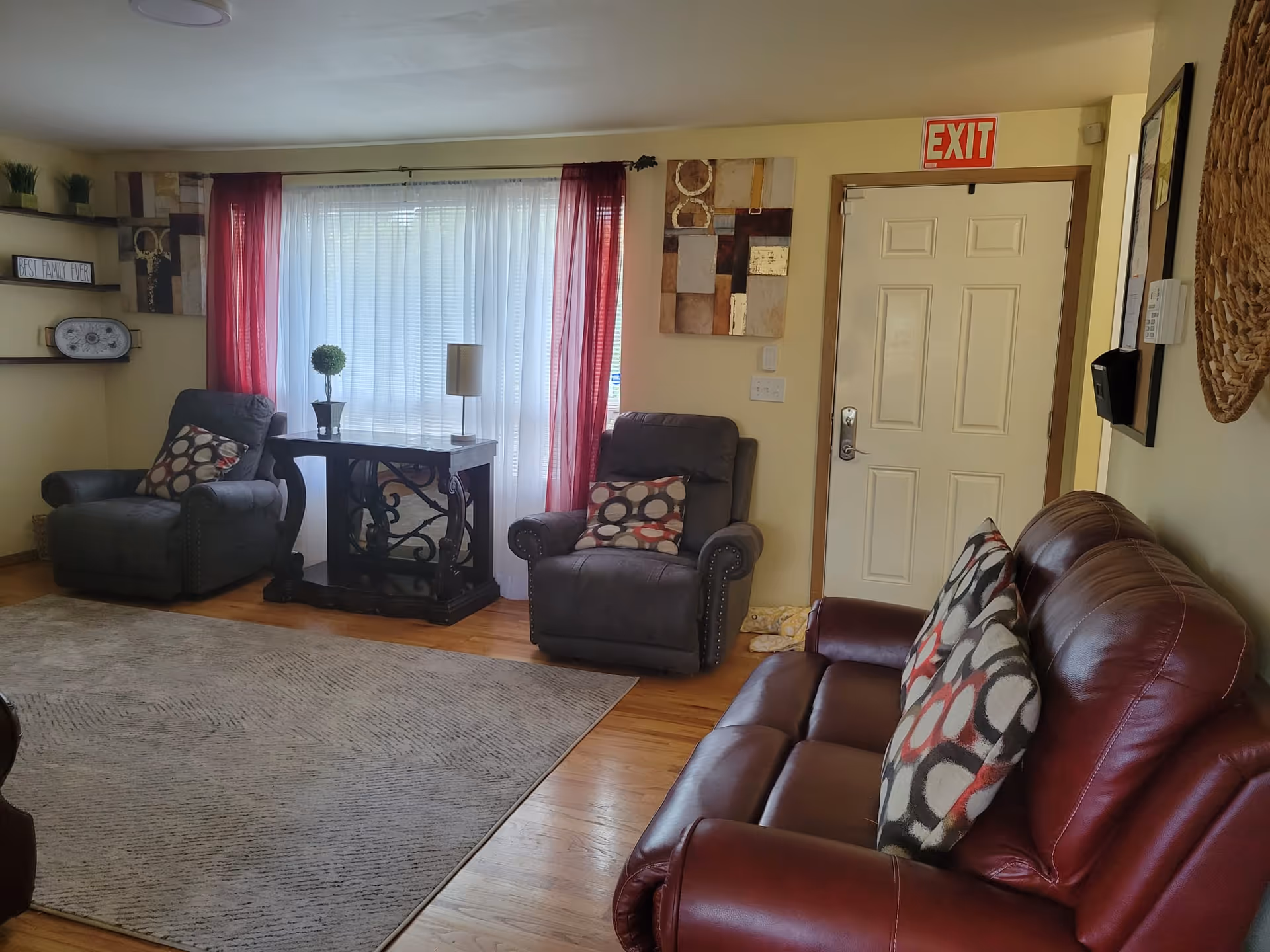 A cozy living room with a red leather sofa and two gray armchairs, each with patterned cushions. There is a window with sheer white curtains and red drapes, a small table with a lamp and a plant, wall art, and an exit door with a red exit sign above it. The floor is wooden with a large area rug.