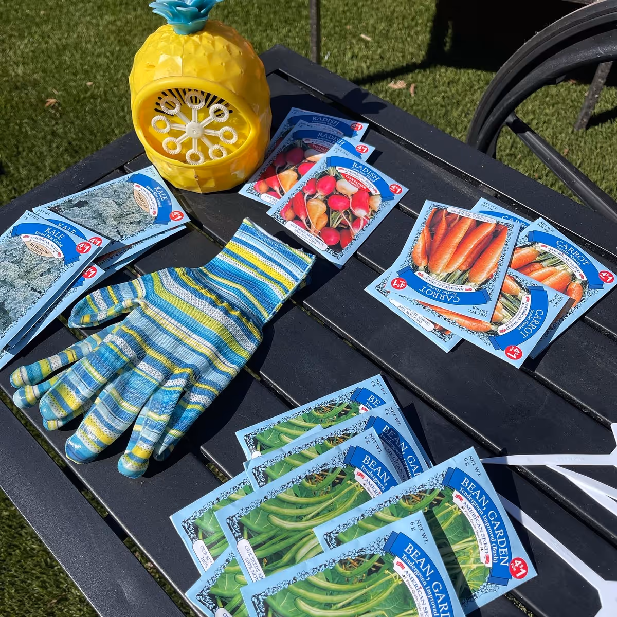 A black outdoor table with several seed packets for kale, radish, carrot, and bean garden laid out. A colorful striped gardening glove and a yellow pineapple-shaped bubble blower are also on the table. The background shows green grass and part of a black metal chair.