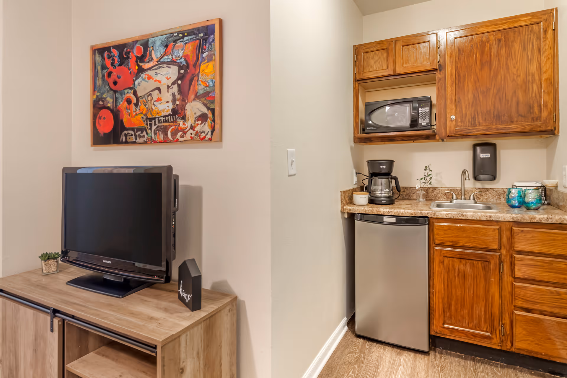 A small kitchenette area with wooden cabinets, a microwave, coffee maker, sink, and mini refrigerator. To the left, there is a wooden TV stand with a flat-screen TV, a small plant, and a decorative block with the word 'home'. Above the TV, a colorful abstract painting is hung on the wall.
