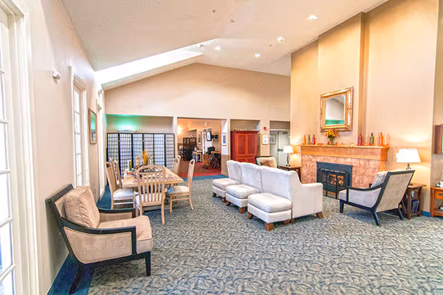 A spacious and well-lit living room area in Green Lake Senior Living Ocean Shores featuring comfortable seating including armchairs and sofas arranged around a fireplace with a mirror above it. There is a dining table with chairs in the background and decorative items on the mantel and side tables. The room has a high ceiling with skylights and carpeted flooring.