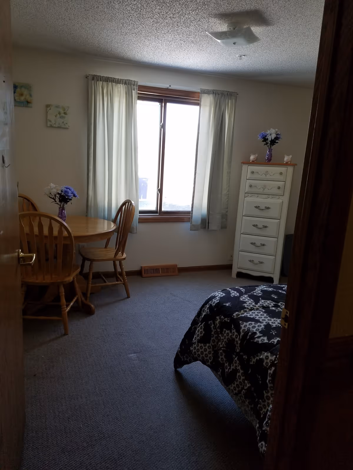 A small bedroom with a window covered by light curtains, a round wooden table with two wooden chairs, a white dresser with decorative items on top, and a bed with a black and white patterned comforter.
