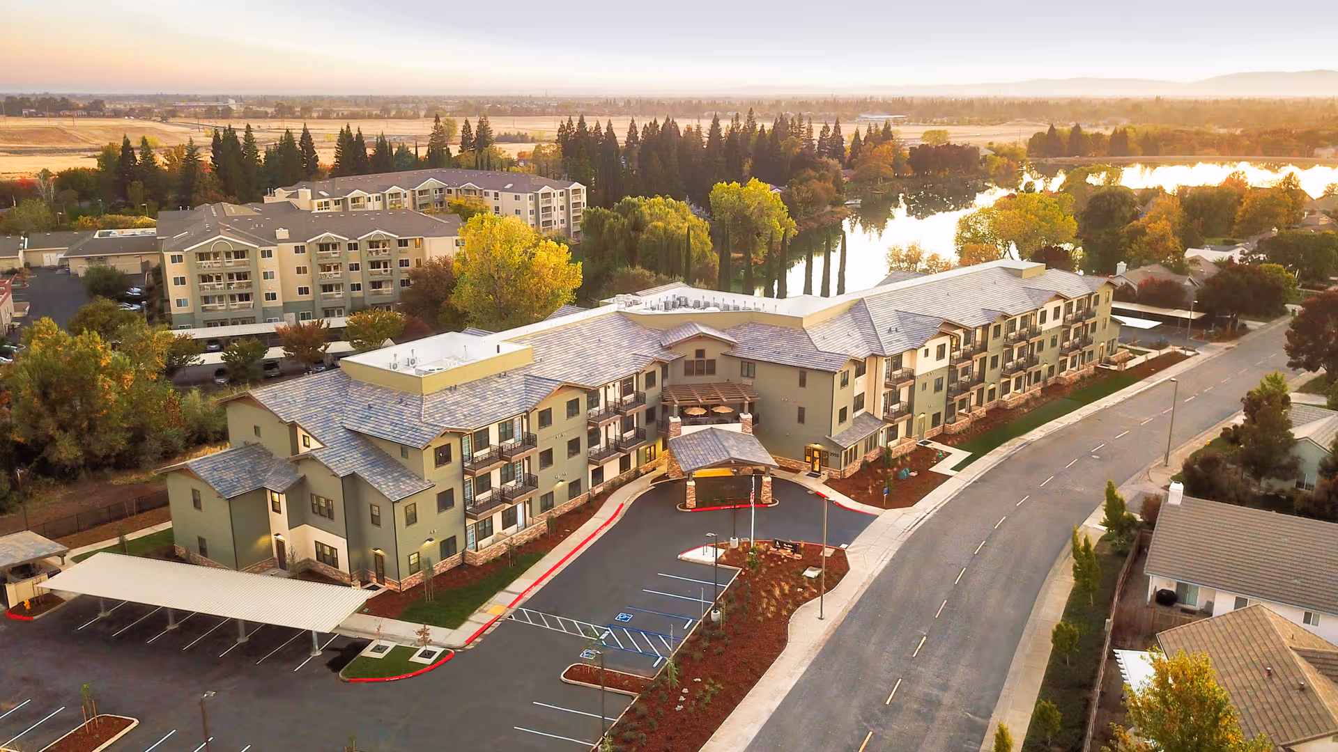 Aerial view of Westmont of Chico senior living facility showing a large multi-story building with balconies, surrounded by trees and landscaping. There is a parking lot with covered parking spaces and a curved road leading to the entrance. In the background, there are more buildings, trees, and a body of water reflecting the sunset.
