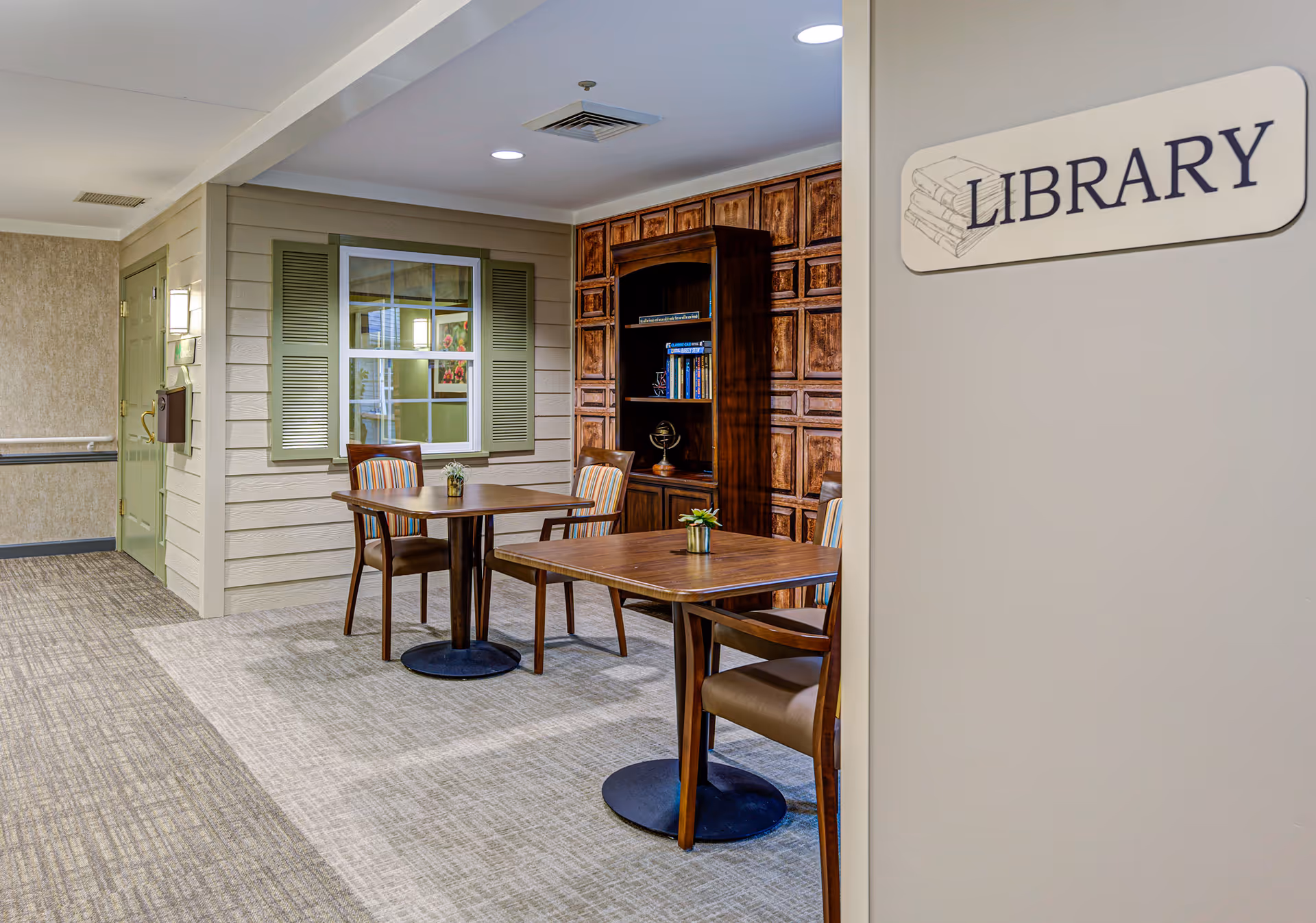 Interior view of a library area in a senior living facility with two wooden tables, each surrounded by chairs. There is a wooden bookshelf with books and decorative items against a wall with a wood panel design. A sign on the wall reads 'LIBRARY'. The floor is carpeted and the walls are painted in neutral tones.