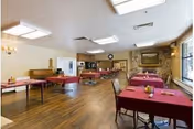 A spacious dining room with multiple tables covered in red tablecloths, each set with condiments. The room has wooden flooring, large windows on one side allowing natural light, ceiling lights, and a clock on the far wall above a doorway. There is a painting on the wall and some chairs around the tables.