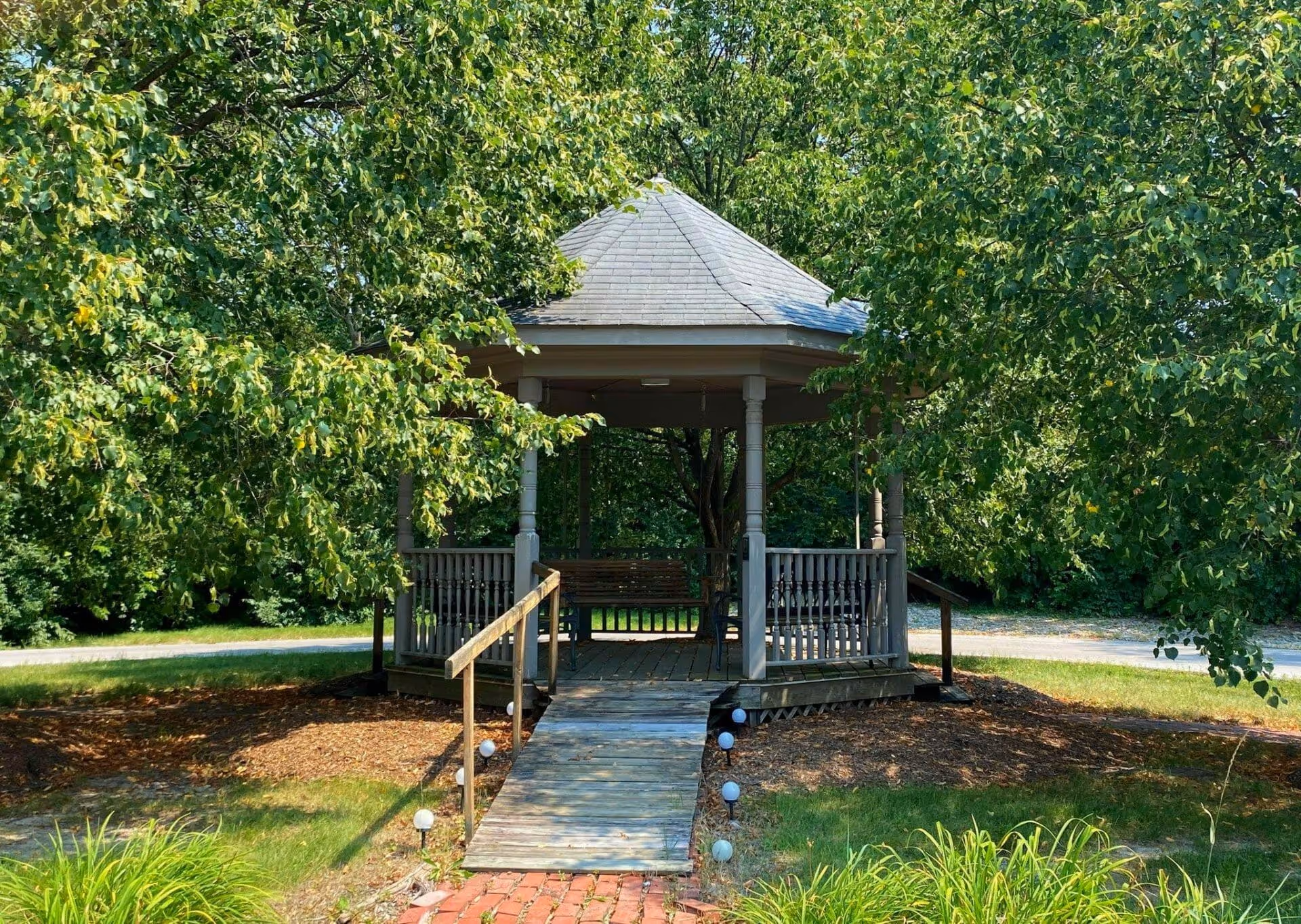 A wooden gazebo with a shingled roof surrounded by lush green trees and grass. The gazebo has a wooden ramp with handrails leading up to it and a bench swing inside. Small round lights line the pathway to the gazebo.