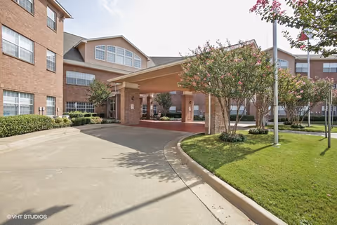 Exterior view of Morada Cedar Hill senior living facility showing a covered entrance with brick pillars, a driveway, manicured green lawn, and flowering trees under a clear sky.