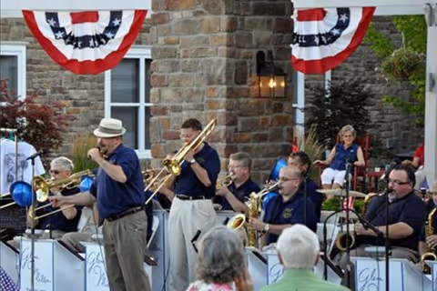 A group of musicians performing outdoors in front of a stone building decorated with red, white, and blue bunting. The band members are playing brass and woodwind instruments, and some audience members are visible in the foreground.