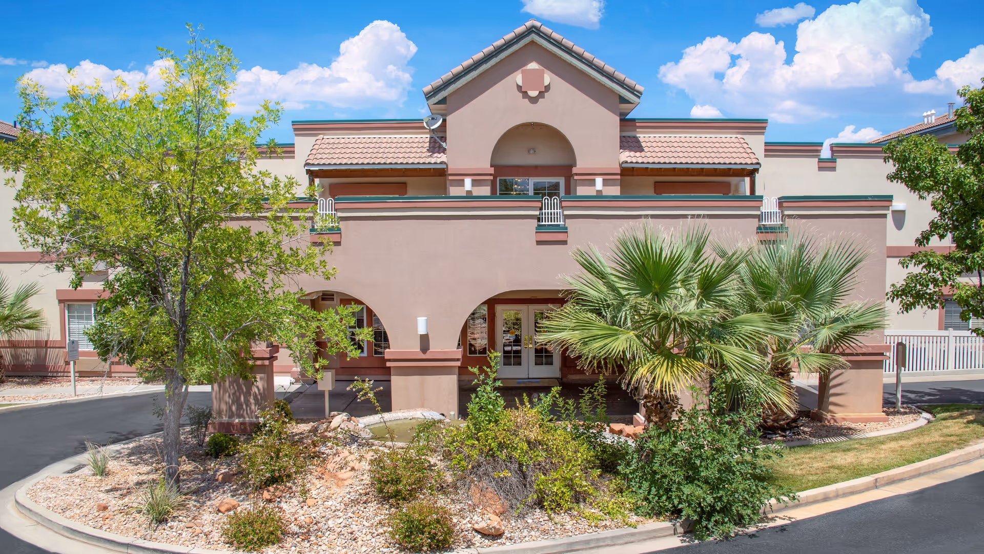 Front exterior view of a two-story senior living facility building with a beige and brown facade, arched entryways, and a landscaped area with trees and shrubs in front under a blue sky with clouds.