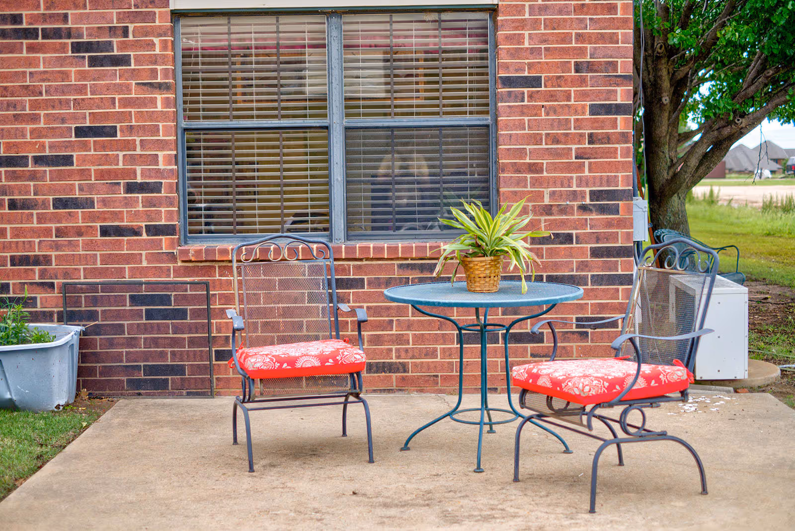 Outdoor patio area with two metal chairs featuring red floral cushions and a round metal table with a potted plant on top, set against a brick wall with a window. There is a tree and some greenery visible in the background.