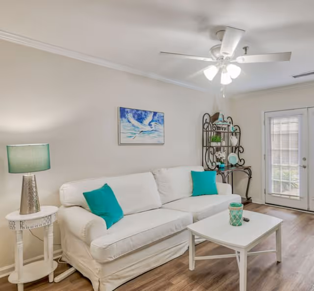 Bright living room featuring a white sofa with turquoise pillows, a white coffee table, side table with a lamp, decorative shelving, and a ceiling fan by glass doors.