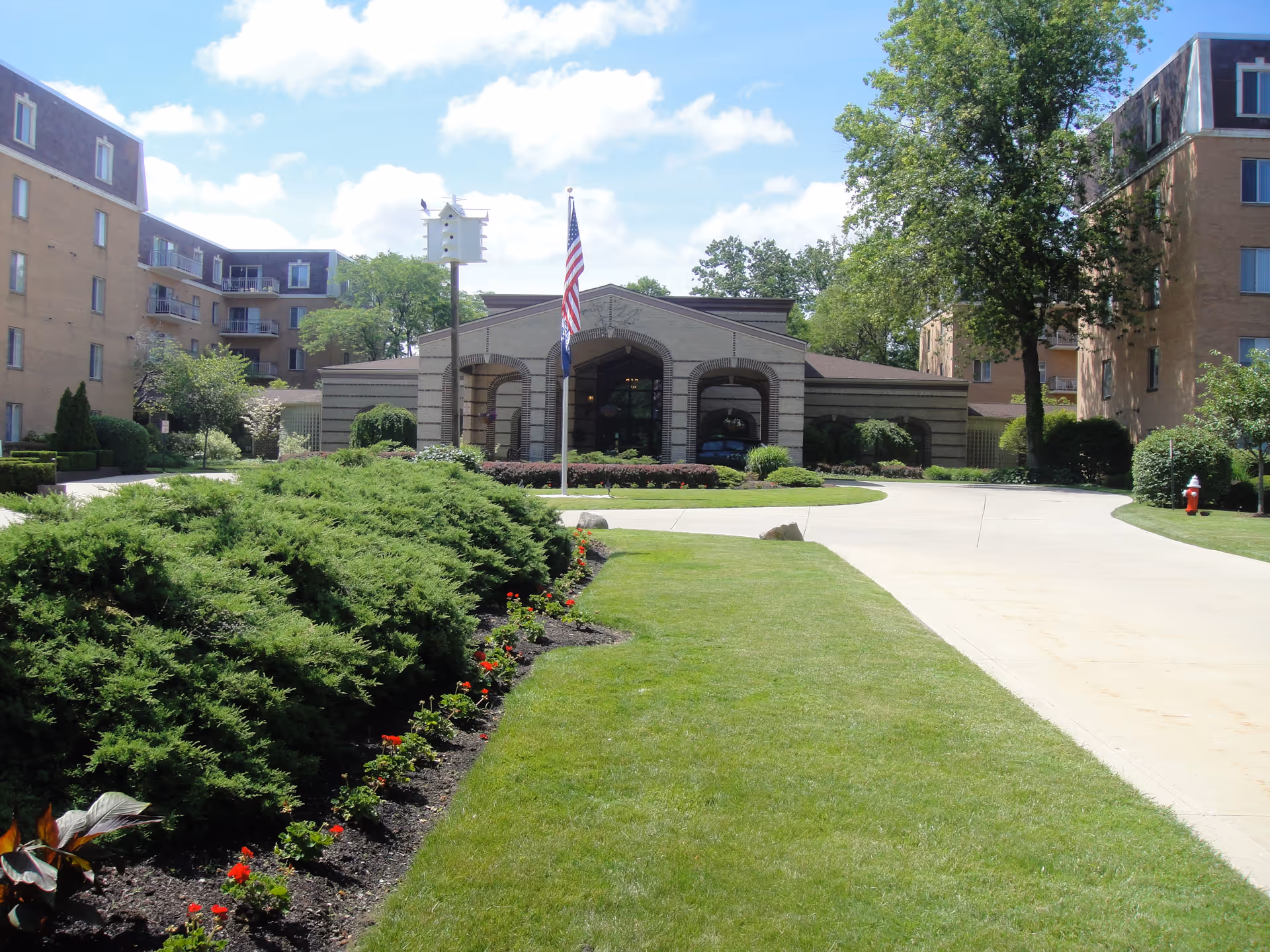 Exterior view of Breakwater at The Normandy senior living facility showing a driveway leading to the main entrance with an American flag and a birdhouse on a pole. The building is surrounded by well-maintained green lawns, bushes, and trees under a partly cloudy sky.