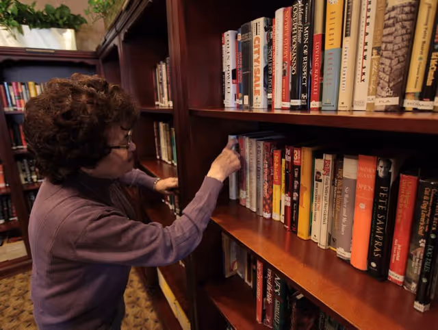 An elderly woman with curly hair and glasses is selecting a book from a wooden bookshelf filled with various books in a library or reading room.