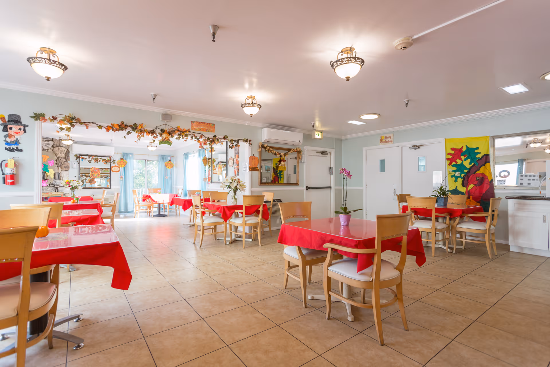 Dining room with multiple square tables draped in red tablecloths, wooden chairs, tiled floor and fall-themed decorations.