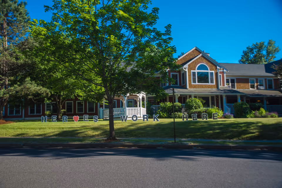 Exterior view of a senior living facility with a large green tree in front and a white gazebo. The building has brown siding with white trim and multiple windows. On the lawn, large letters spell out 'HEROES WORK HERE' with a red heart symbol replacing the letter O in HEROES.