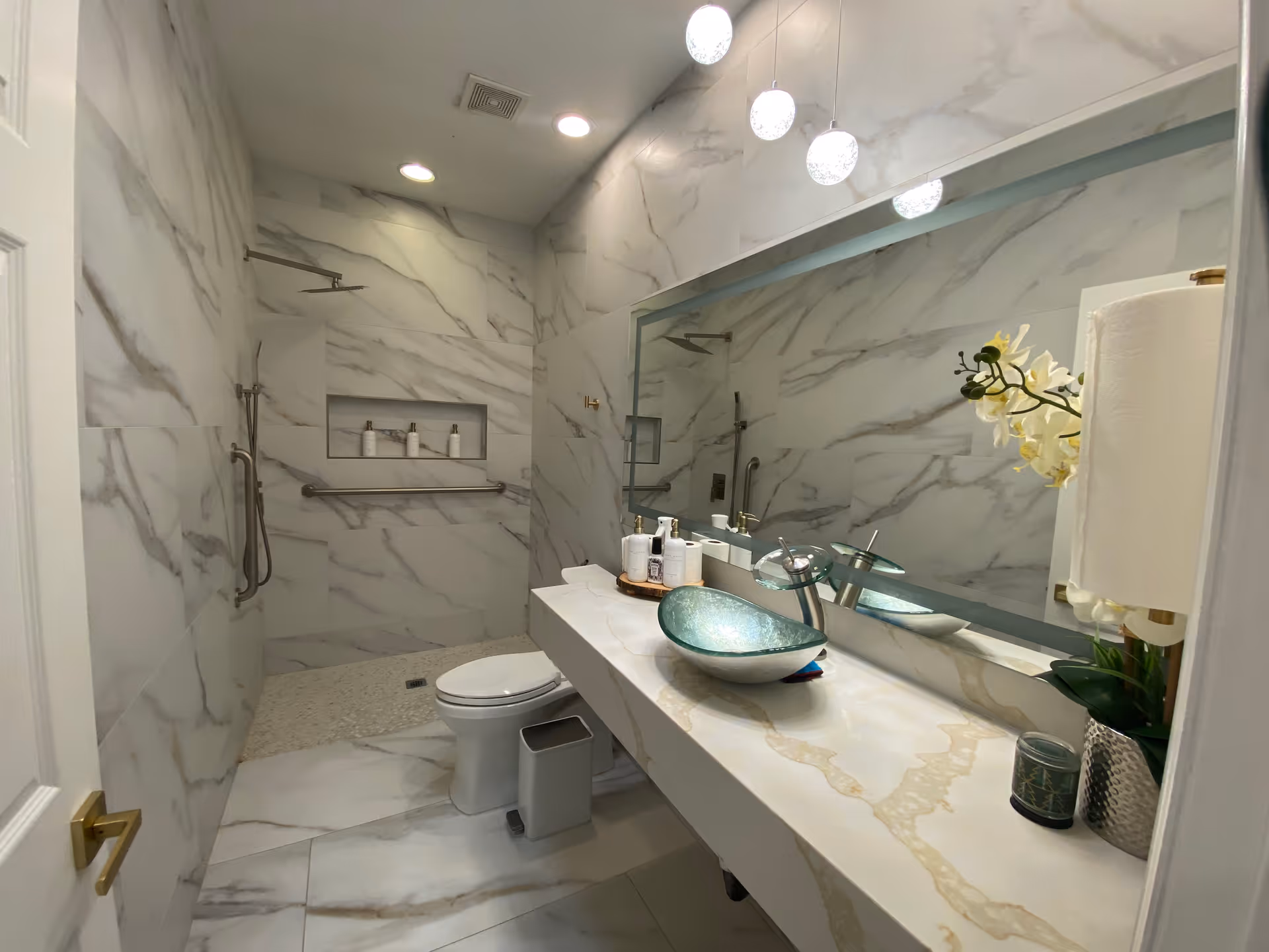 Modern marble bathroom with a walk-in shower, wall-mounted grab bars, a glass vessel sink on a long countertop, toilet, and large mirror.