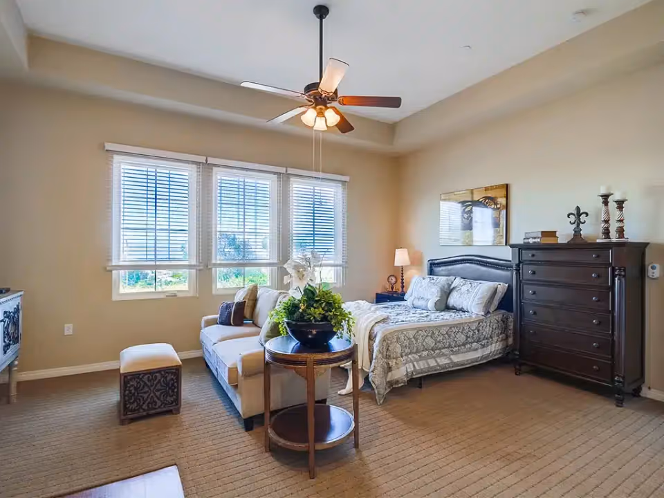 Spacious furnished bedroom with a bed, nightstand, dresser, sofa, side table with plant, ceiling fan, and three large windows with blinds.