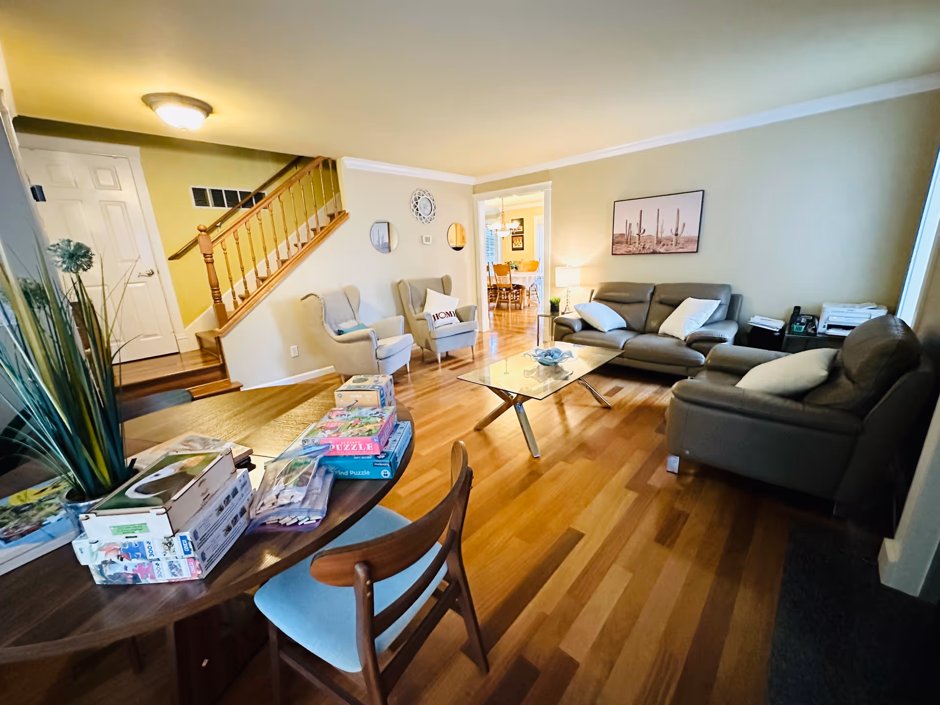 A cozy living room with wooden flooring, featuring two gray armchairs with a pillow on one, a gray sofa with white pillows, a glass coffee table, and a wooden table with puzzles and games on it. There is a staircase with wooden railing, a door, and a wall clock and mirrors on the wall. The room is warmly lit with a ceiling light and a table lamp, and a framed picture of cacti hangs on the wall.