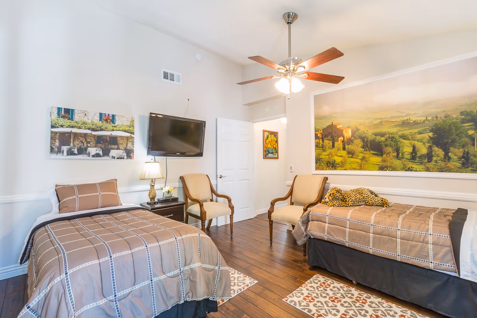 Bright shared bedroom with two twin beds, chairs, a wall-mounted TV, ceiling fan, and large landscape artwork.