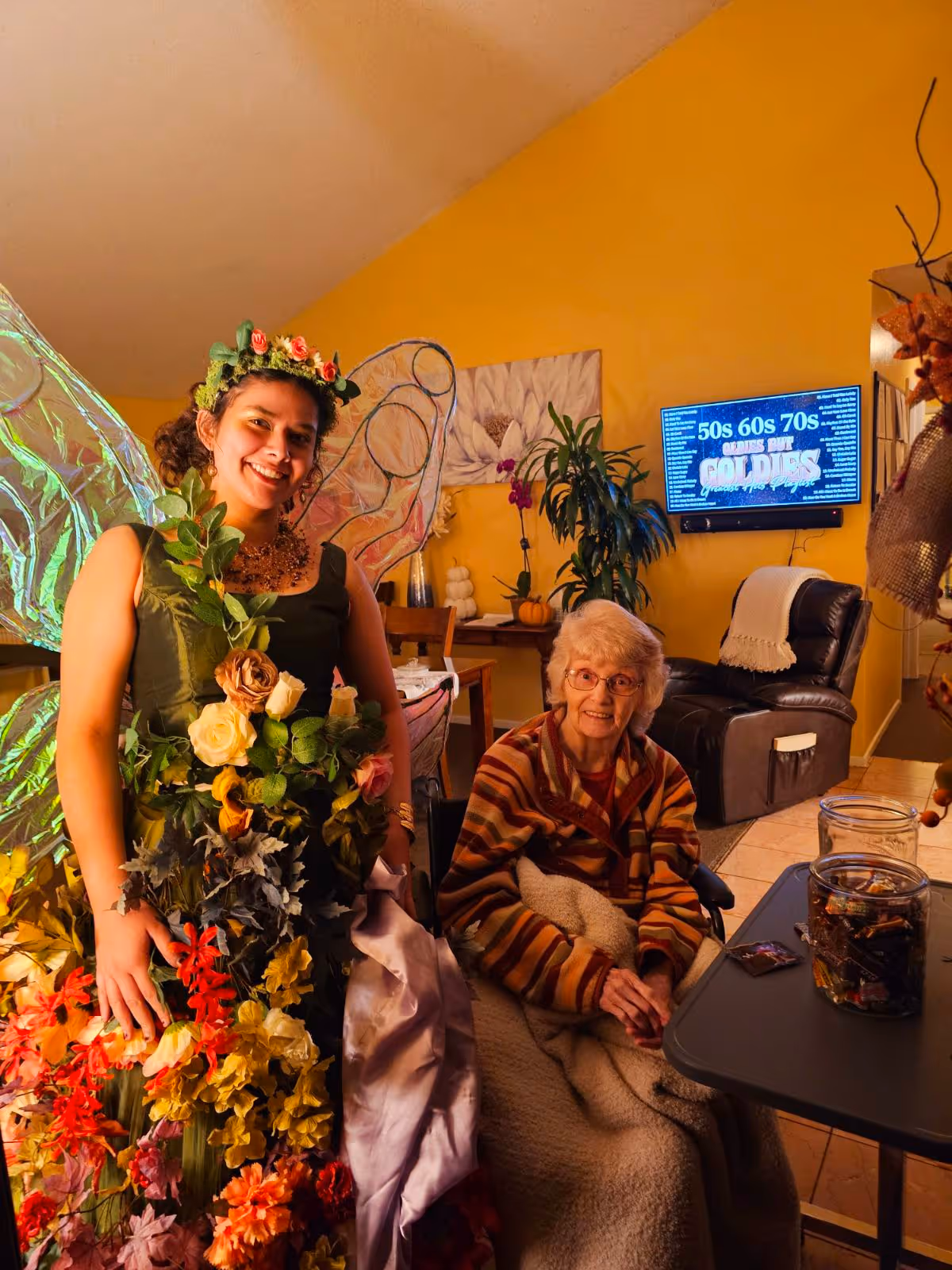 A young woman dressed as a fairy with colorful flowers and green wings stands next to an elderly woman sitting in a wheelchair covered with a blanket in a warmly lit living room with yellow walls. Behind them is a TV screen displaying text about the 50s, 60s, and 70s, and there are plants and furniture in the background.