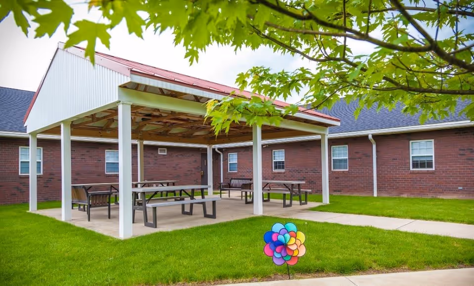 Outdoor covered seating area with picnic tables and benches on a concrete slab, surrounded by green grass and a brick building in the background. A colorful pinwheel decoration is placed in the grass near the sidewalk, and green tree leaves frame the top of the image.