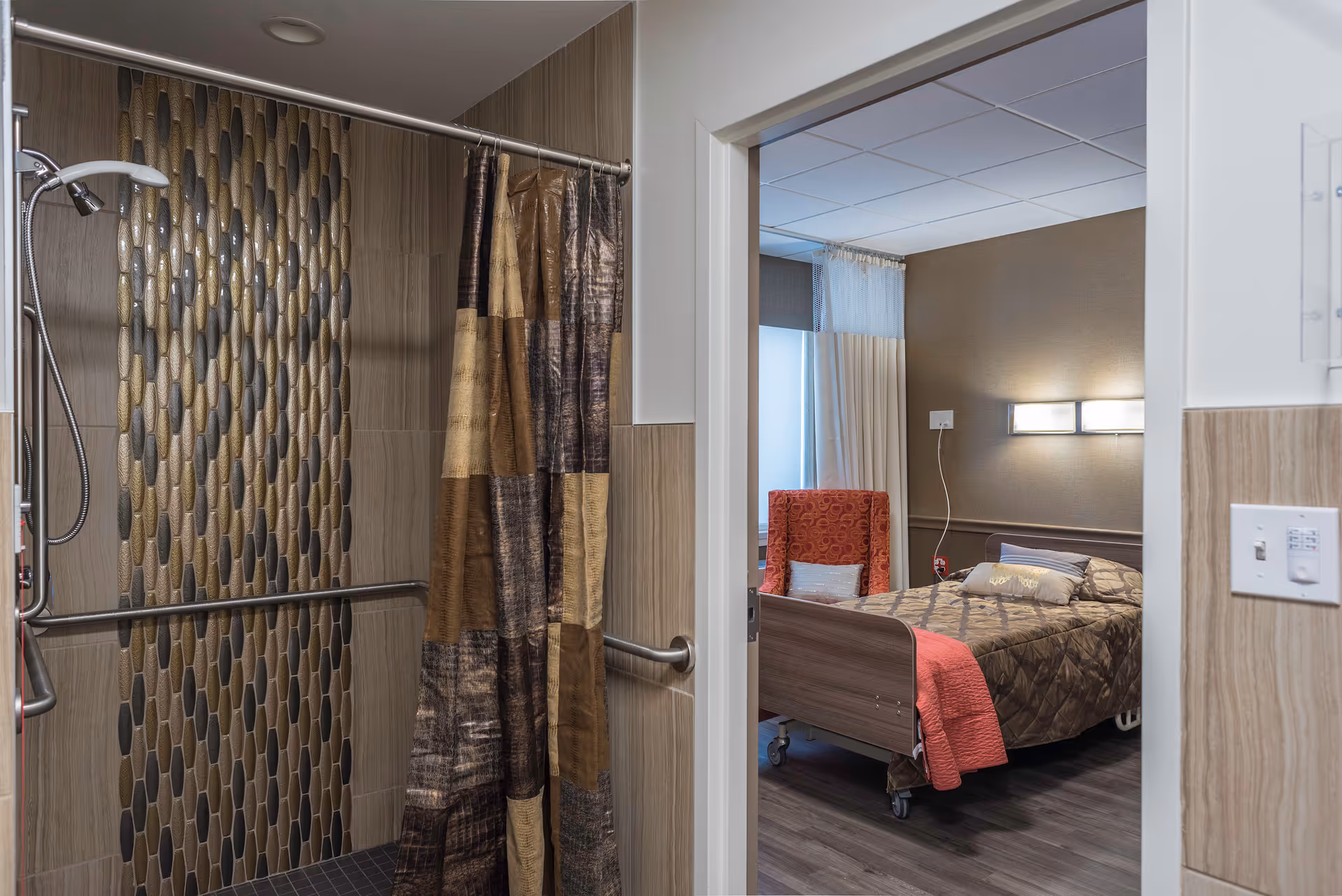 View of a bathroom with a shower area featuring a patterned tile wall and a brown shower curtain, adjacent to a bedroom with a hospital-style bed, a red patterned armchair, and a window with curtains.