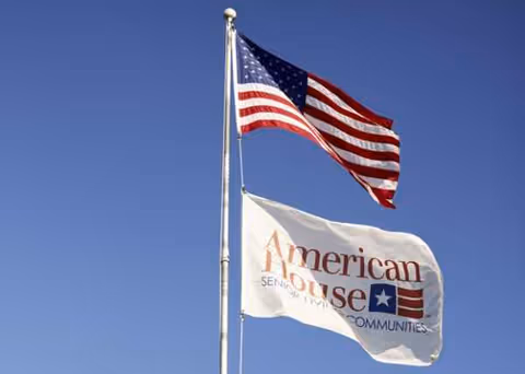 Two flags, American flag and American House flag, fly on tall poles against a blue sky.