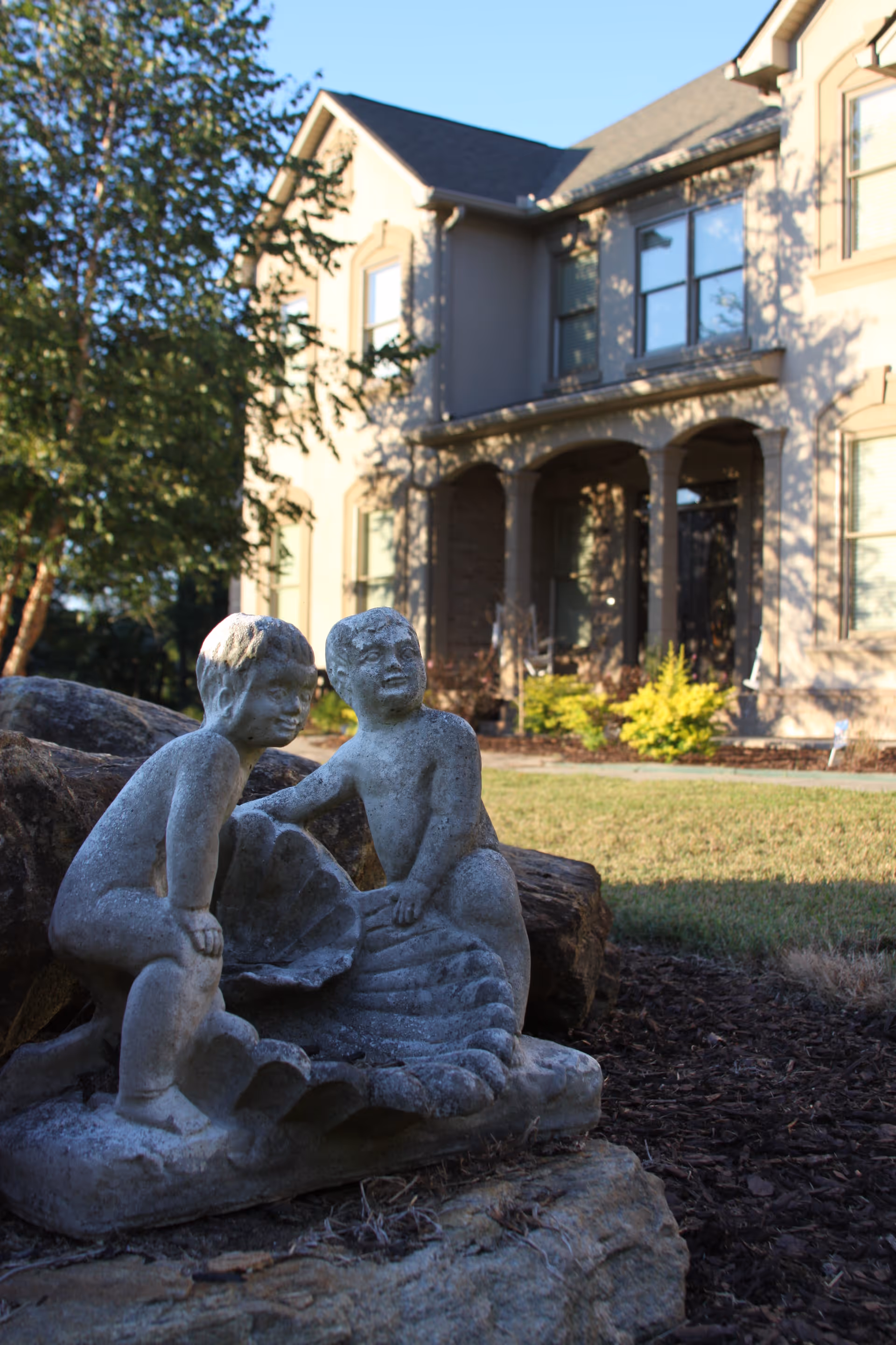 Stone statue of two children in a shell on the lawn with a two-story house in the background.