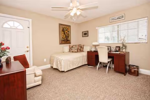 A beige bedroom in a senior living suite with bed, desk, and seating.