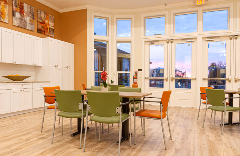 A bright and spacious dining area with light wood flooring, white cabinets along one wall, and several tables surrounded by green and orange chairs. Large windows and glass doors let in natural light and offer a view of the outdoors during sunset. Decorative artwork is displayed above the cabinets.