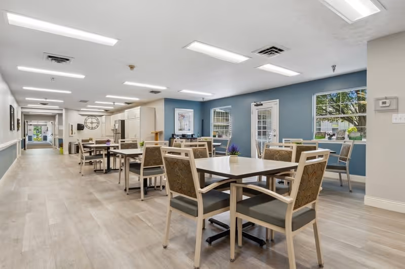 A bright and spacious dining area in a senior living facility with multiple square tables and chairs arranged neatly. The room has light wood flooring, blue and white walls, large windows letting in natural light, and fluorescent ceiling lights. A hallway extends from the dining area, and there is a door leading outside.