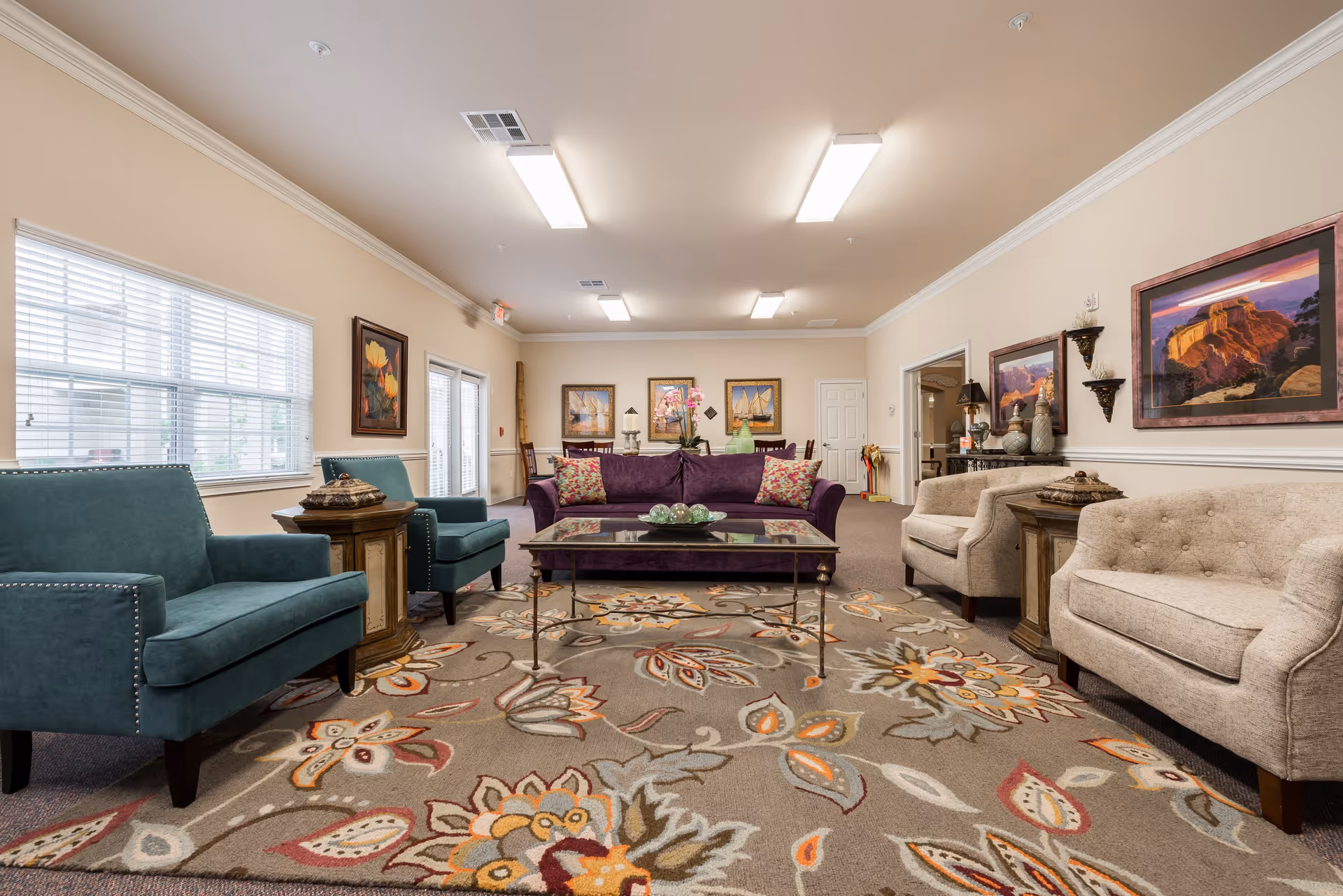 A spacious senior living facility common area with a large floral patterned rug, a purple sofa with colorful cushions, two teal armchairs, and two beige armchairs. The room has beige walls adorned with framed artwork and decorative wall sconces. There are large windows with white blinds on the left side, and ceiling lights illuminate the room.