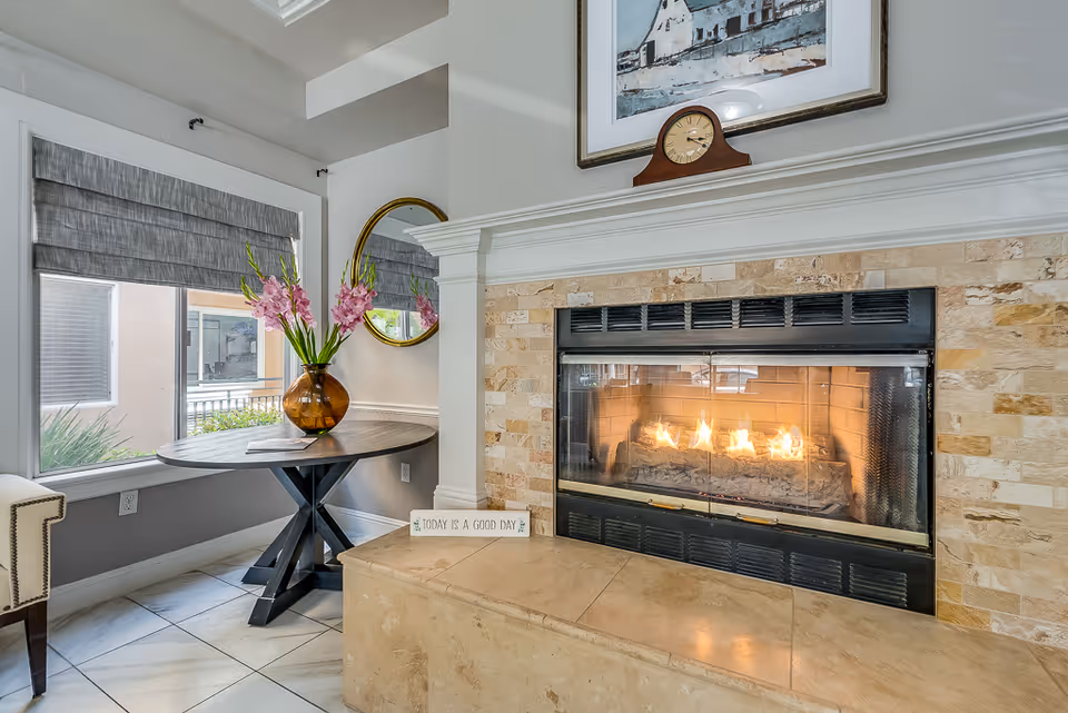 Cozy interior corner featuring a lit fireplace with a stone surround and a mantel displaying a clock and a framed painting. Next to the fireplace is a round wooden table with a vase of pink flowers, a round mirror on the wall, and a window with gray Roman shades.