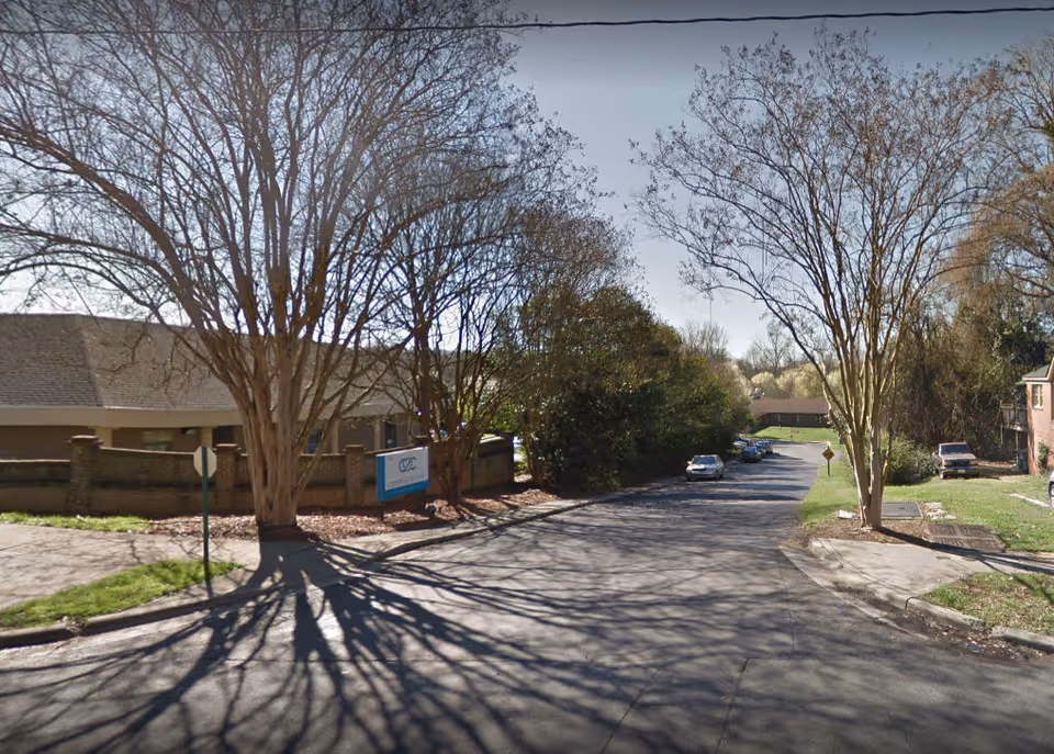 Street view of a residential area with a building partially visible on the left side behind a brick wall and leafless trees casting shadows on the road. Several parked cars are visible along the street, and there is a sign near the building entrance.