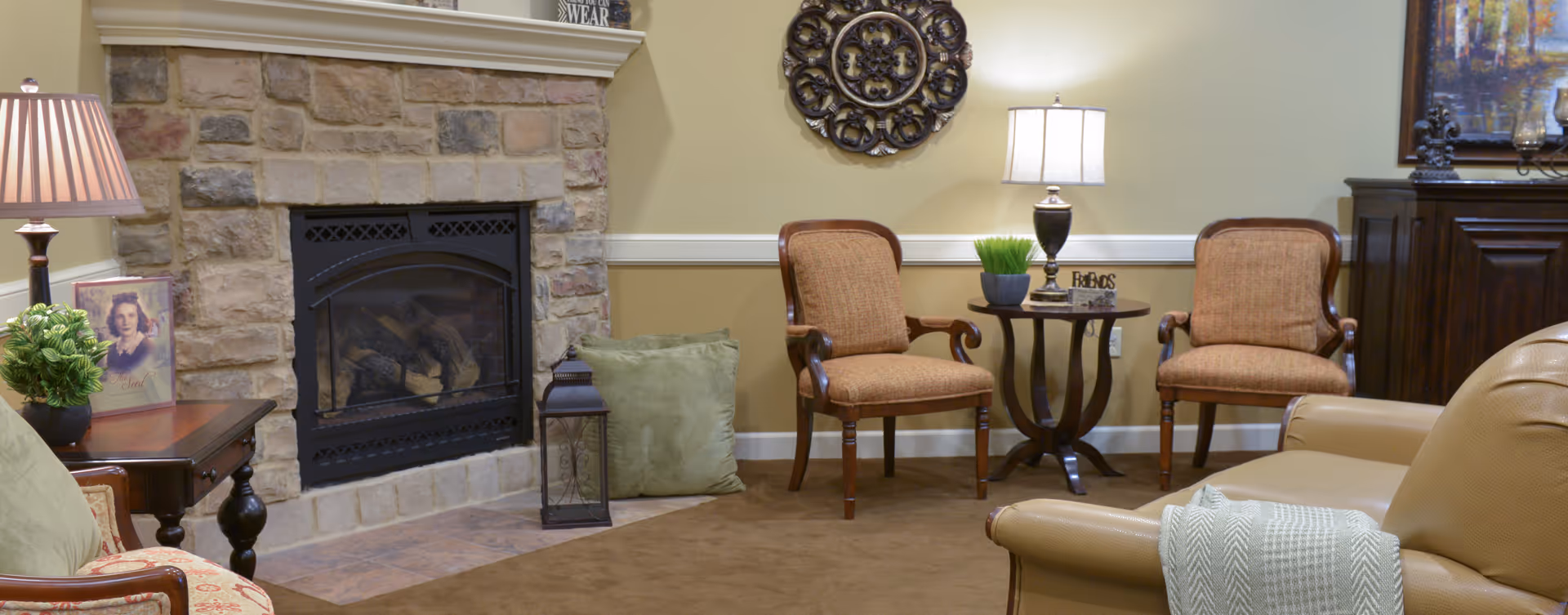 A cozy living room area with a stone fireplace, two upholstered chairs, a small round wooden table with a lamp and decorative items, a side table with a lamp and a framed photo, and a beige leather armchair with a green throw blanket.