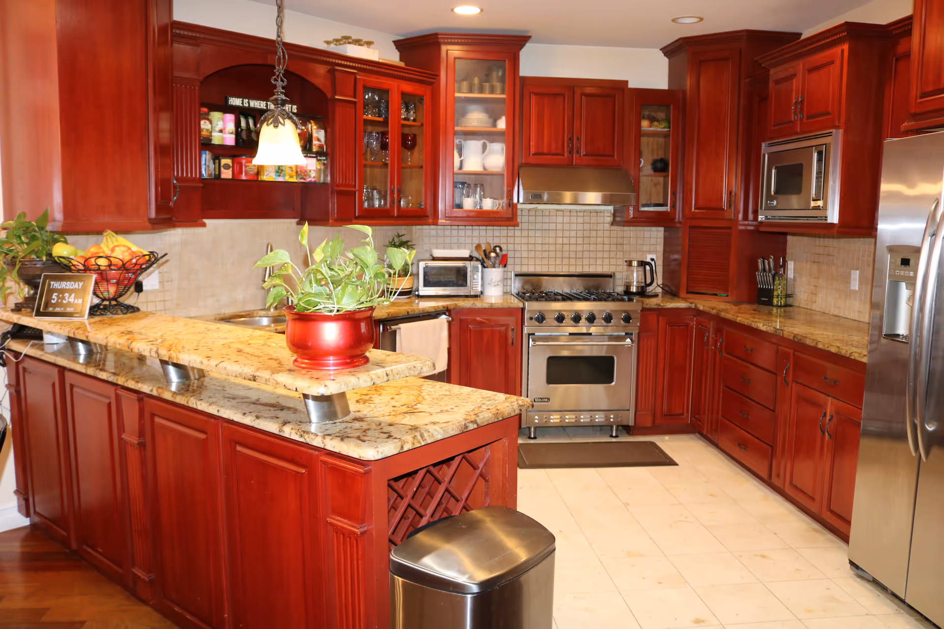 A spacious kitchen with rich wooden cabinets, granite countertops, and stainless steel appliances including a stove, microwave, and refrigerator. There is a small plant in a red pot on the counter, a fruit basket, and various kitchen utensils and glassware visible. The floor is tiled and the kitchen is well-lit with ceiling lights and a hanging lamp.