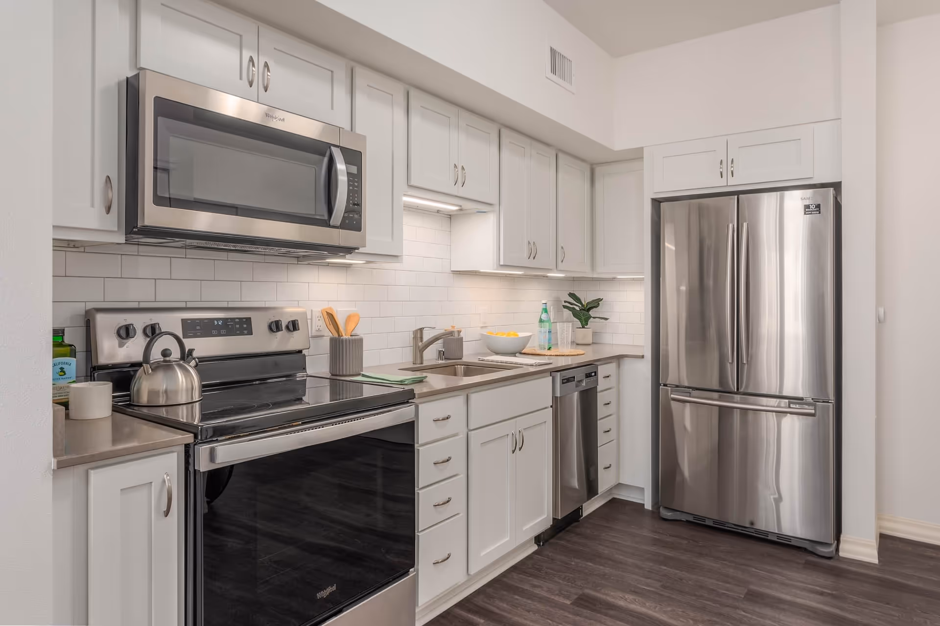 Modern kitchen with white cabinets, stainless steel appliances including a microwave, stove, dishwasher, and refrigerator. The countertop has a sink, a bowl of lemons, a plant, and some glasses. The backsplash is white subway tile and the floor is dark wood.