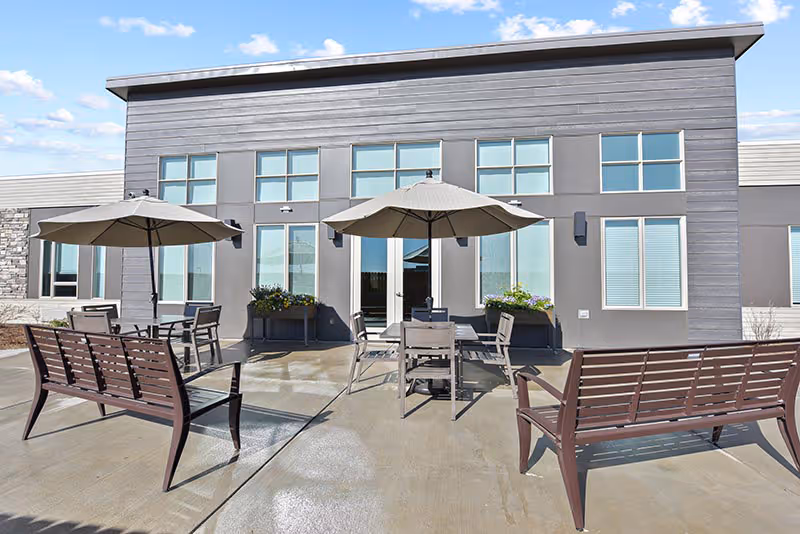 Outdoor patio area with two brown benches and two tables each shaded by a large umbrella, situated in front of a modern building with multiple windows and flower planters.